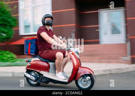 Il ragazzo di consegna indossa un uniforme rossa su scooter con scatola di cibo isotermico che guida velocemente. Servizio espresso di consegna di cibo da caffè e ristoranti Foto Stock