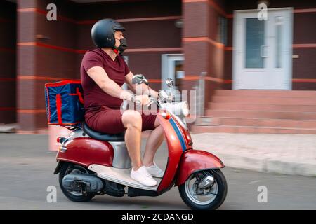 Il ragazzo di consegna indossa un uniforme rossa su scooter con scatola di cibo isotermico che guida velocemente. Servizio espresso di consegna di cibo da caffè e ristoranti Foto Stock