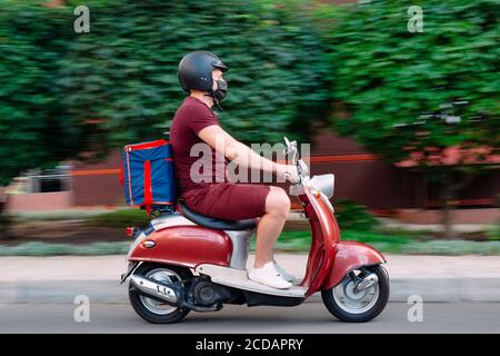 Il ragazzo di consegna indossa un uniforme rossa su scooter con scatola di cibo isotermico che guida velocemente. Servizio espresso di consegna di cibo da caffè e ristoranti Foto Stock