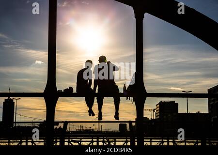 Persone retroilluminate non identificabili che godono del sole serale seduto su un ponte di acciaio sopra i binari ferroviari chiamato Haccurbrücke. Monaco, Germania Foto Stock