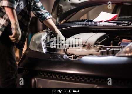 un giovane meccanico maschile in una camicia e guanti controlla il livello dell'olio motore in una vettura nera Foto Stock
