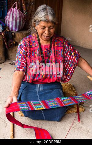 Una donna maya Cakchiquel più anziana in abito tradizionale esamina il colorato tessuto faja o cintura che ha appena fatto a Santa Catarina Palopo, Guatemala. Foto Stock