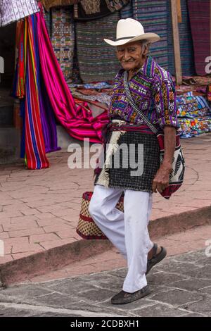 Un uomo Maya più vecchio di Tzutujil in abito tradizionale cammina lungo la strada a Santiago Atitlan, Guatemala. Indossa un pesante rodillera intrecciato intorno alla sua wa Foto Stock