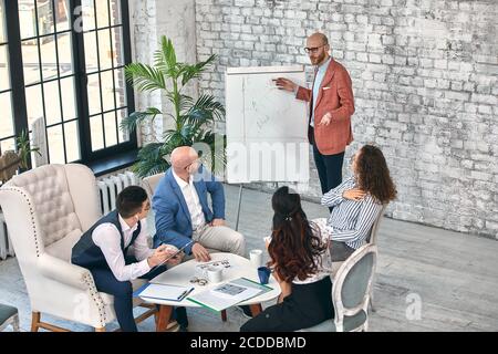 Uomo d'affari sicuro che scrive sulle lavagne a fogli mobili presentazione nuovo progetto in sala riunioni alla riunione aziendale. Un auditor mentore serio parla con i partner Foto Stock