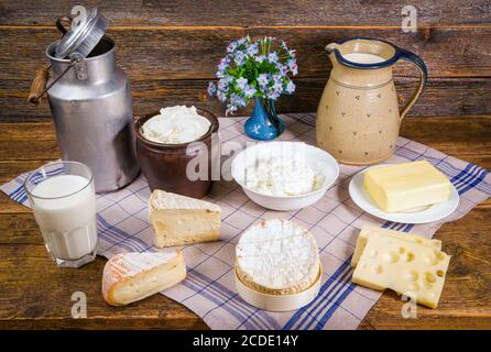 Latte in una caraffa e un bicchiere, diversi tipi di formaggio, panna montata, cagliata e un pezzo di burro in un ambiente rustico su un tavolo di legno con cucina Foto Stock