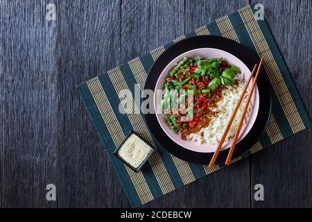 Maiale tritato su riso con fagioli verdi cosparsi di semi di sesamo, peperoncino rosso servito su una ciotola con bastoncini su un vecchio fondo di legno, piatto, c Foto Stock