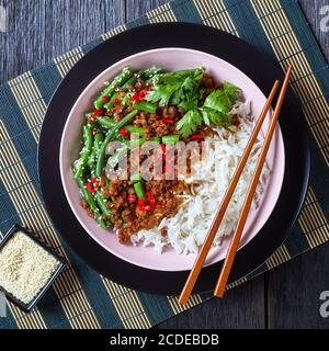 Piatto di maiale tritato al basilico thailandese kra pao con riso a grani lunghi e fagioli verdi cosparsi di semi di sesamo, peperoncino rosso servito su un recipiente rosa con bastoncini Foto Stock