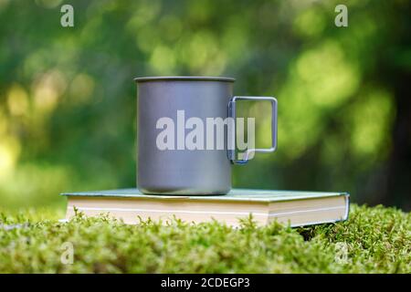 Tazza di titanio su un libro nella foresta. Lo sfondo di Fern e muss è sfocato. Foto Stock