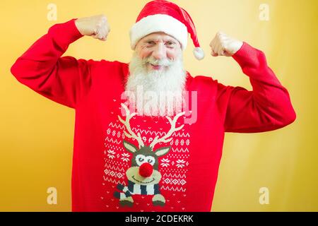 Uomo anziano sopportato in una festa di Natale con sfondo giallo. Felice persona matura che indossa cappelli babbo natale. Vacanze e gioioso stile di vita anziano c Foto Stock