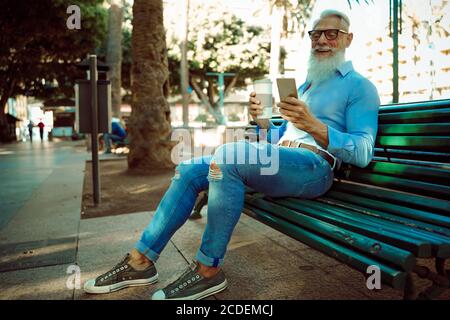 Uomo anziano sorridente che si rilassa nel parco. Maschio maturo che si diverte con nuove tendenze tecnologiche e bere caffè. Foto Stock