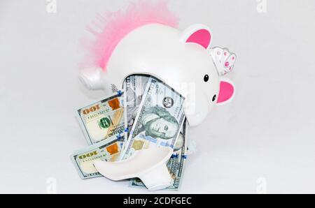 Le banconote da un centinaio di dollari stanno versando fuori da una banca piggy di capretti di ceramica bianca rotta con le orecchie rosa e il tuto rosa su uno sfondo bianco. Foto Stock