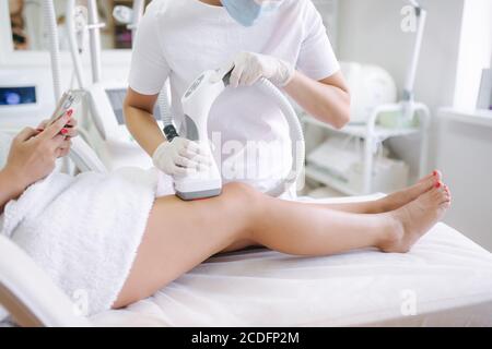 Beutiful bussines donna che usa il telefono mentre cosmetologist in guanti bianchi che esegue la procedura di sollevamento a radiofrequenza sullo stomaco. Primo piano Foto Stock