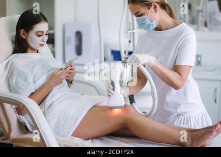 Beutiful bussines donna con maschera facciale bianca utilizzando il telefono mentre cosmetologo in guanti bianchi che esegue la procedura di sollevamento a radiofrequenza su Foto Stock