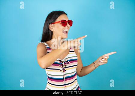 Giovane bella donna che indossa costume da bagno e occhiali da sole su blu isolato sfondo sorpreso e guardando il lato che punta con due mani e. dita a. Foto Stock