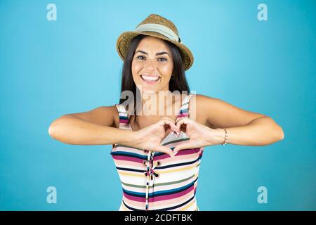 Giovane bella donna che indossa costume da bagno e cappello su blu isolato sfondo sorridente in amore mostrando il simbolo del cuore e la forma con mani Foto Stock