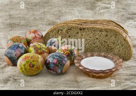 Molti freschi rurale di pasqua uova decorate giacenti in un wattled Piccolo cesto vicino al pane fresco e saltcellar su un panno da un roug Foto Stock