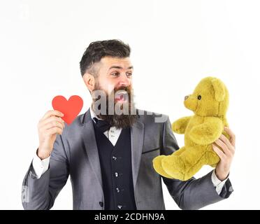 L'uomo in costume con il volto sorridente tiene un orso peluche. Gentleman con barba e baffi tiene il cuore rosso o carta valentine, isolato su sfondo bianco. Concetto di giorno di San Valentino. L'uomo si prepara per la data. Foto Stock