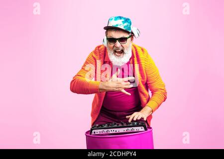 DJ. Ritratto di uomo hipster anziano in occhiali alla moda isolato su sfondo rosa studio. Concetto di stile di vita per anziani con tecnologia e gioia. Colori alla moda, per sempre giovani. CopySpace per il tuo annuncio. Foto Stock