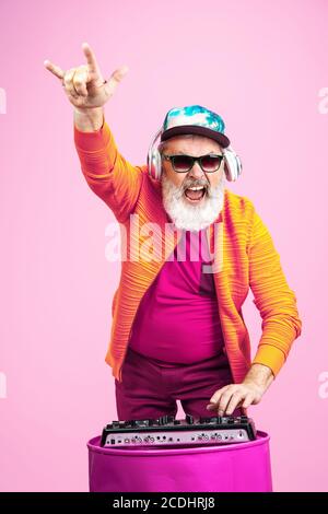 Party drive. Ritratto di uomo hipster anziano in occhiali alla moda isolato su sfondo rosa studio. Concetto di stile di vita per anziani con tecnologia e gioia. Colori alla moda, per sempre giovani. CopySpace per il tuo annuncio. Foto Stock