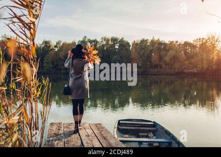 Donna che cammina sul molo del lago in barca ammirando il paesaggio autunnale che tiene le foglie. Attività della stagione autunnale Foto Stock