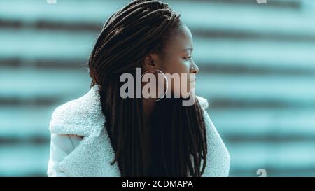 Profilo di una giovane adorabile femmina nera con grandi dreadlock, orecchini rotondi, e piercing nel naso, sta guardando da parte mentre sta in piedi all'aperto Foto Stock