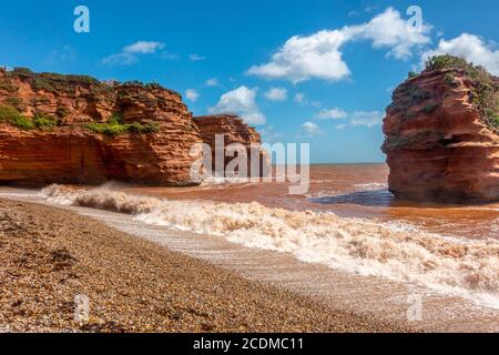 Le onde rotolano sulla spiaggia a Ladram Bay nel Devon del sud, Inghilterra. Le scogliere di arenaria si sono erose nel tempo per lasciare una pila indipendente Foto Stock