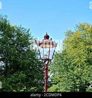 Versione moderna replica elettrica di una lampada da strada a gas in ferro battuto vittoriano ornato, sullo sfondo di alberi e un cielo blu. Foto Stock