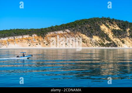 Piccola barca che si snoda lungo la costa occidentale dell'isola di Fraser con dune colorate sullo sfondo Foto Stock