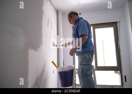 Pittore di casa anziano reale in piedi su scala che dipinge una parete grigia. Vero lavoratore sporco. Concetto di professione del pittore della casa. Foto Stock