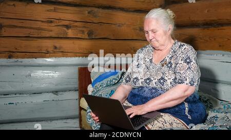 la donna anziana con i capelli grigi tiene un laptop in grembo seduto su un letto. Foto Stock