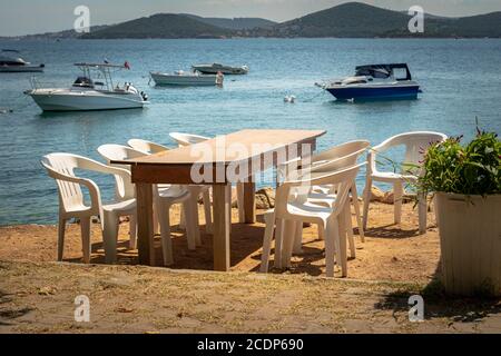 tavolo in legno e sedie in plastica sulla spiaggia che si affaccia sul mare dove sono ancorate piccole barche, caldo giorno d'estate, nessuno. Foto Stock