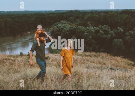 I genitori della famiglia camminano con la madre esterna del bambino e il padre che trasporta bambino figlia sulle spalle sano stile di vita rurale natura fiume e. vista sulla foresta Foto Stock