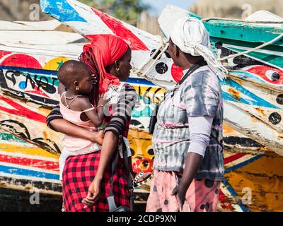 Nianing, Senegal - 24 gennaio 2019: Donna senegalese e la chiocciera sullo sfondo di colorate barche da pesca in legno in piedi sulla spiaggia di sabbia in Senegal. Foto Stock