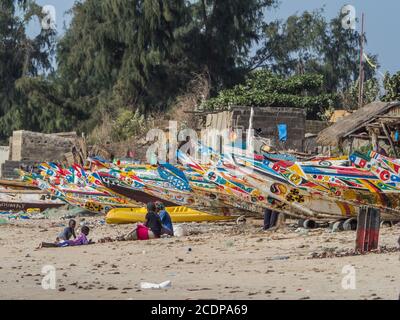 Nianing, Senegal - 24 gennaio 2019: Famiglia africana che siede vicino alle colorate barche da pesca in legno in piedi sulla spiaggia di sabbia in Senegal. Africa Foto Stock