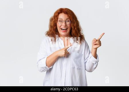 Ritratto di piacevole sorridente donna di mezza età con capelli rossi, indossare occhiali e blusa mostrando pubblicità, cliente di azienda consiglia prodotto o. Foto Stock