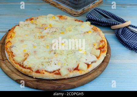 Pizza tradizionale su un vassoio di legno sul tavolo. Pizza rotonda con pollo, mais, ananas, formaggio. Cottura fatta in casa. Forcella, coltello, sfondo blu Foto Stock