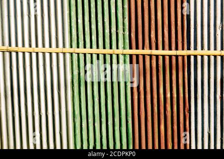 La parete corrugata verniciata multicolore conferisce tonalità orizzontali, strisce. Un bastone orizzontale di bambù sta tenendo insieme la recinzione arrugginita. Foto Stock