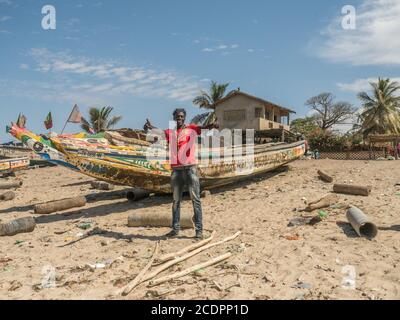 Nianing, Senegal - 24 gennaio 2019: Sorridente senegalese sullo sfondo di colorate barche da pesca in legno in piedi sulla spiaggia di sabbia in Senegal. AFR Foto Stock