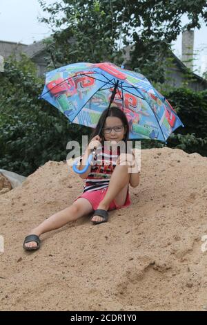 ragazza sotto ombrello luminoso giocare sotto la pioggia in giardino nell'ultimo giorno d'estate in vacanza Foto Stock
