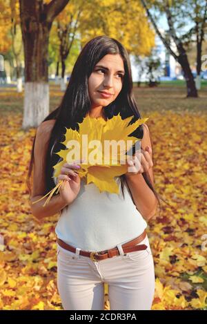 Bella ragazza in parco autunno tenendo una foglia d'acero in mano, sorridendo sul suo volto, in jeans bianchi e una blusa, su uno sfondo di alberi e voi Foto Stock