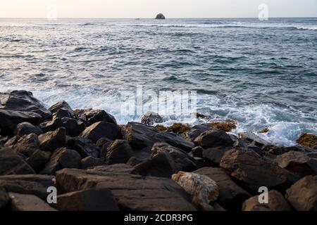 una barriera rocciosa intemperata da un'azione ondulata inarrestabile e un enorme masso in mare sullo sfondo lontano Foto Stock