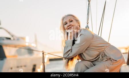 Ritratto di una donna caucasica matura con lunghi capelli grigi guardando direttamente alla macchina fotografica mentre si siede su una barca a vela a. tramonto Foto Stock
