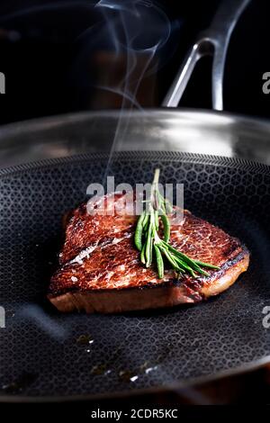 Pezzo di bistecca di manzo preparazione sulla padella scura con olio ed erbe, fuoco selettivo Foto Stock