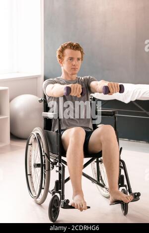 Giovane uomo in activewear seduto in sedia a rotelle e allenandosi con manubri Foto Stock