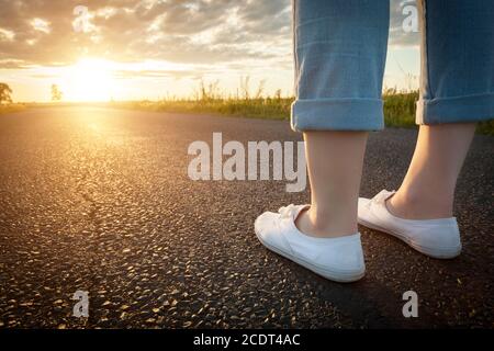 Donna in sneakers bianche in piedi su strada asfaltata verso il sole. Concetti di viaggio, libertà. Foto Stock