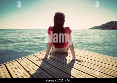 Giovane donna che riposa sul molo guardando il mare calmo nella soleggiata giornata estiva. Foto Stock