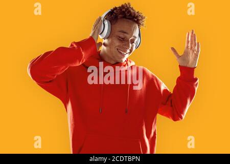 Stile libero. Ragazzo mulatto in felpa con cappuccio e cuffie in piedi isolati sul giallo ascolto di musica danzante gioioso al buio Foto Stock