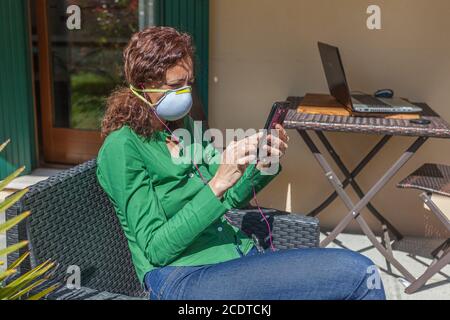 Donna seduta in un giardino con maschera antivirus che guarda cellulare Foto Stock