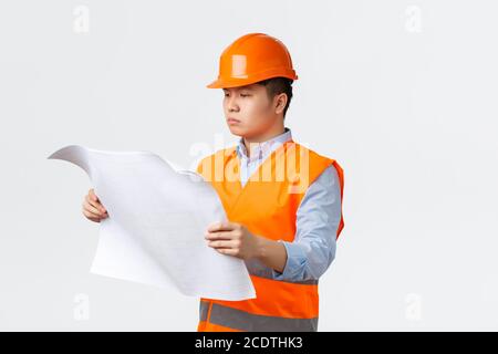 Concetto di settore edilizio e lavoratori industriali. Responsabile della costruzione asiatica serio-osservante, architetto che studia i progetti, guardando il layout con Foto Stock