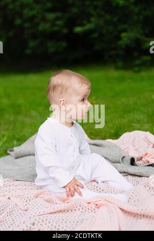 il bambino piccolo in vestiti bianchi siede sulla coperta lavorata a maglia dentro parcheggiare su erba verde Foto Stock